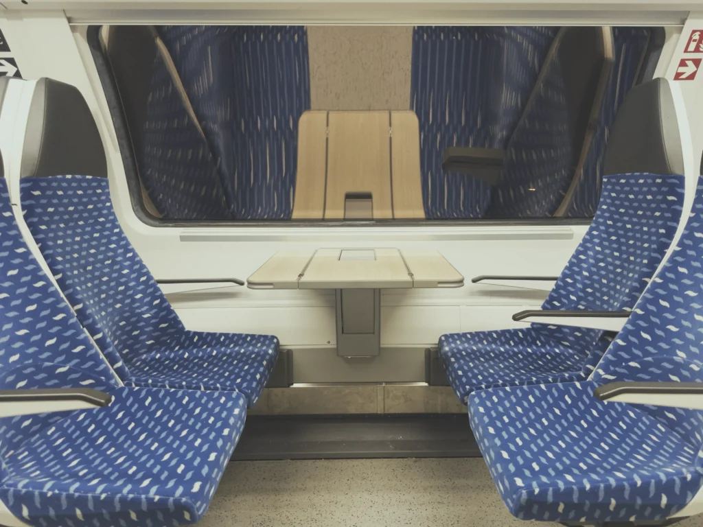 Das Bild zeigt ein Vierer-Sitzabteil in einem Zug. Die Sitze sind blau mit weißem Muster, dazwischen ein kleiner Tisch. Im Hintergrund ist ein Fenster mit Spiegelungen von den Sitzen zu sehen.