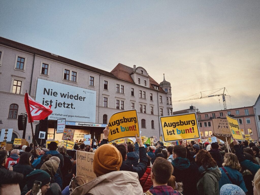 Menschenmenge demonstriert, hält Plakate mit Texten "Nie wieder ist jetzt" und "Augsburg ist bunt!" vor einem historischen Gebäude in einer städtischen Umgebung.